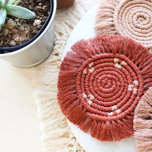 May include: Close-up of handmade macrame coasters in shades of terracotta and beige. The coasters have a spiral design with fringed edges. A potted succulent plant is visible in the background, along with a cream-colored macrame placemat.