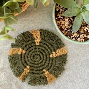 May include: A handmade macrame coaster in olive green and tan. The circular coaster features a spiral design with tan accents and fringe. The coaster is next to a potted succulent and a small plant in a woven basket.