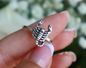 May include: A silver-colored ring featuring a detailed scorpion design. The scorpion has a textured body and raised claws. The ring is shown on a finger with a blurred floral background.