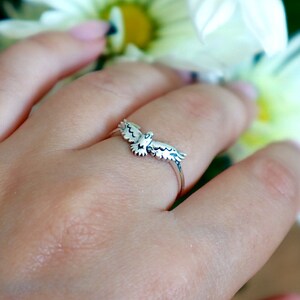 K&ouml;nnte beinhalten: Ein silberner Ring mit einem detaillierten Vogeldesign mit ausgebreiteten Fl&uuml;geln. Der Ring wird an einem Finger gezeigt, mit einem verschwommenen floralen Hintergrund. Die Fl&uuml;gel des Vogels sind aufwendig detailliert.
