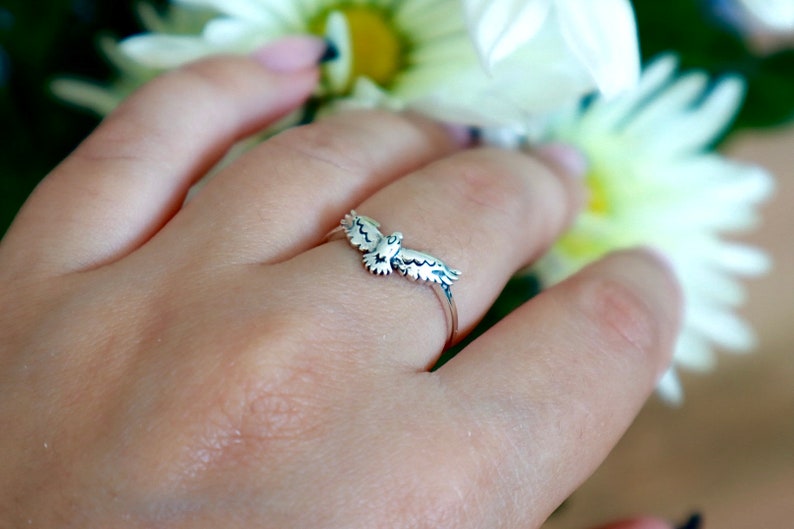 K&ouml;nnte beinhalten: Ein silberner Ring mit einem detaillierten Vogeldesign mit ausgebreiteten Fl&uuml;geln. Der Ring wird an einem Finger gezeigt, mit einem verschwommenen floralen Hintergrund. Die Fl&uuml;gel des Vogels sind aufwendig detailliert.
