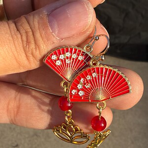 May include: A pair of gold-tone drop earrings featuring red enamel fan-shaped charms decorated with small white floral patterns. Each earring includes a round red bead and a gold-tone lotus flower charm suspended from the fan by a metal link.