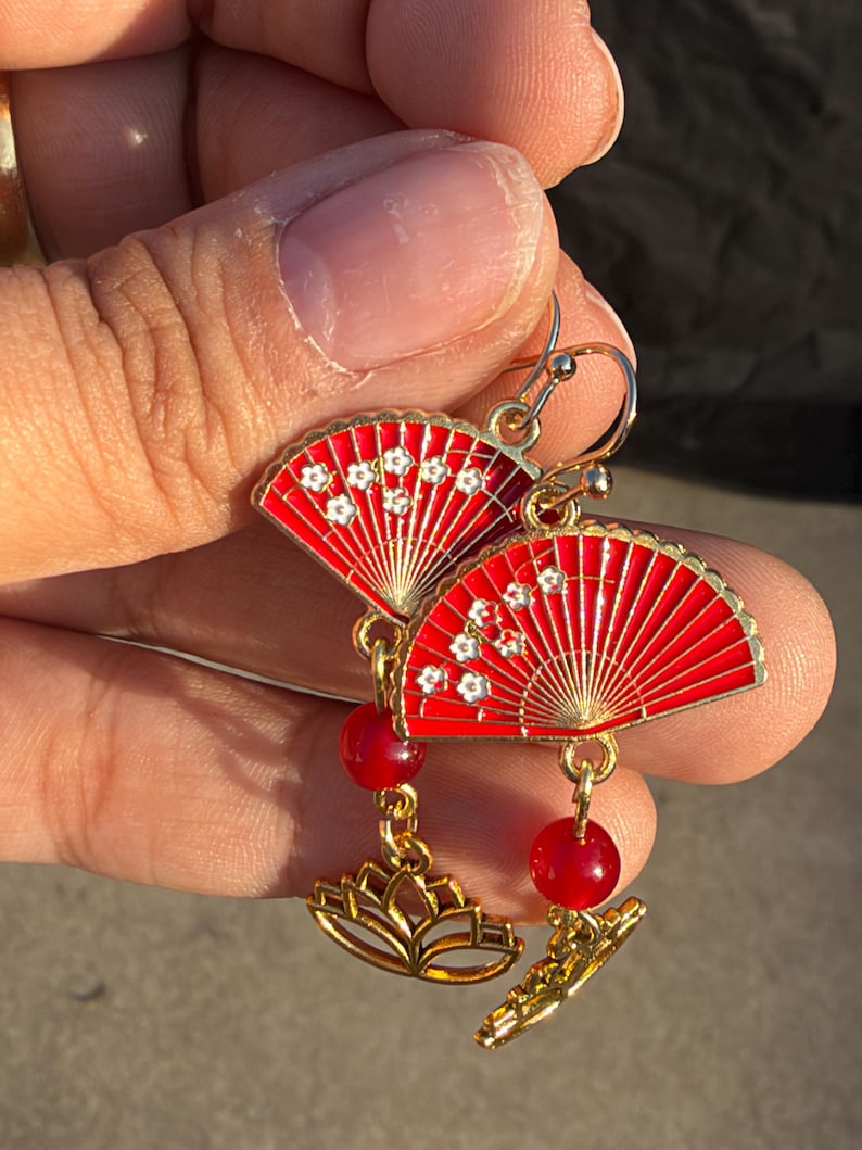 May include: A pair of gold-tone drop earrings featuring red enamel fan-shaped charms decorated with small white floral patterns. Each earring includes a round red bead and a gold-tone lotus flower charm suspended from the fan by a metal link.