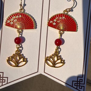 May include: A pair of gold-tone dangle earrings featuring a red fan-shaped charm with small white floral accents, a single round red bead, and a gold-tone lotus flower charm at the bottom, suspended from gold-tone earring hooks.