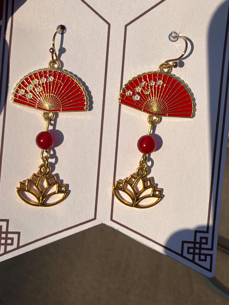 May include: A pair of gold-tone dangle earrings featuring a red fan-shaped charm with small white floral accents, a single round red bead, and a gold-tone lotus flower charm at the bottom, suspended from gold-tone earring hooks.
