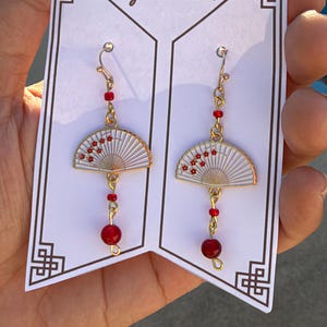 May include: A pair of gold-toned dangle earrings featuring white fan-shaped charms with red floral accents. Each earring includes a small red bead above the fan and a larger red bead at the bottom, suspended from a gold hook on a display card.