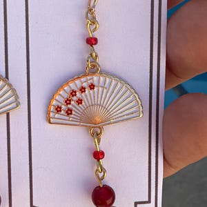 May include: A gold-toned drop earring featuring a white folding fan charm with red floral accents. The earring includes a small red bead above the fan and a larger red bead dangling below, all connected by gold-toned metal links and a hook wire.