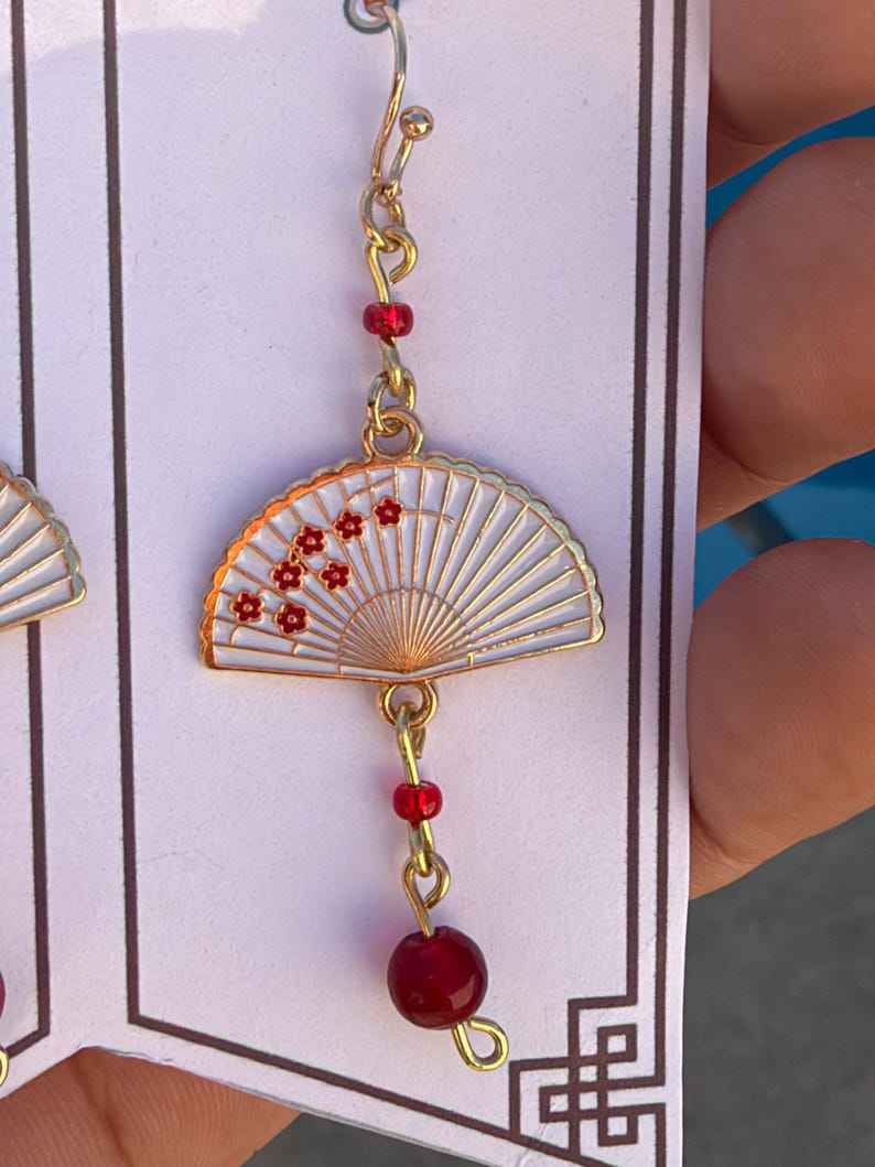 May include: A gold-toned drop earring featuring a white folding fan charm with red floral accents. The earring includes a small red bead above the fan and a larger red bead dangling below, all connected by gold-toned metal links and a hook wire.