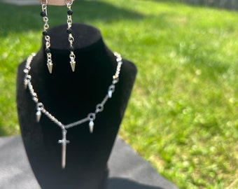 May include: A silver necklace with a sword pendant and small spikes hanging from the chain. Matching earrings with black beads and silver spikes.