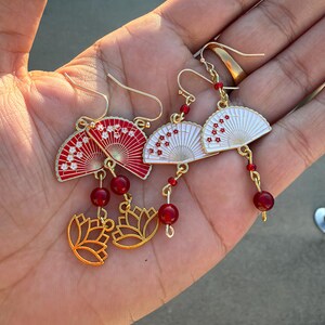 May include: Two pairs of gold-tone dangle earrings featuring fan-shaped charms. One pair has red fans with white floral accents, and the other has white fans with red floral accents. Each earring includes a red bead and a gold-tone lotus charm.