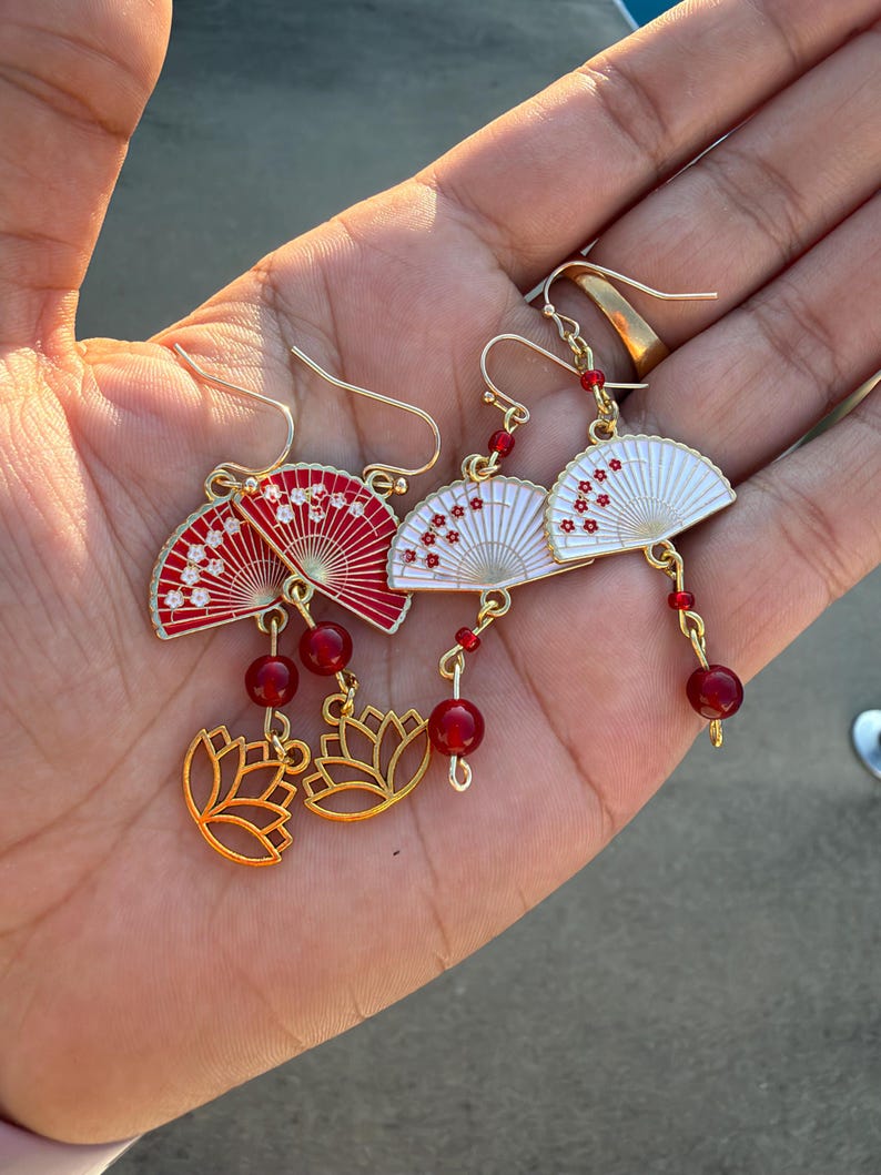 May include: Two pairs of gold-tone dangle earrings featuring fan-shaped charms. One pair has red fans with white floral accents, and the other has white fans with red floral accents. Each earring includes a red bead and a gold-tone lotus charm.