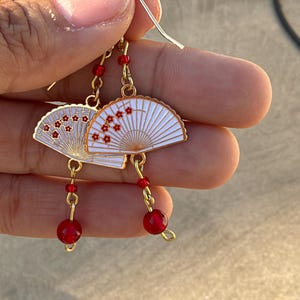 May include: A pair of gold-toned dangle earrings featuring white fan-shaped charms decorated with small red floral patterns. Each earring is accented with a single small red bead above the fan and a larger red bead hanging from the bottom.