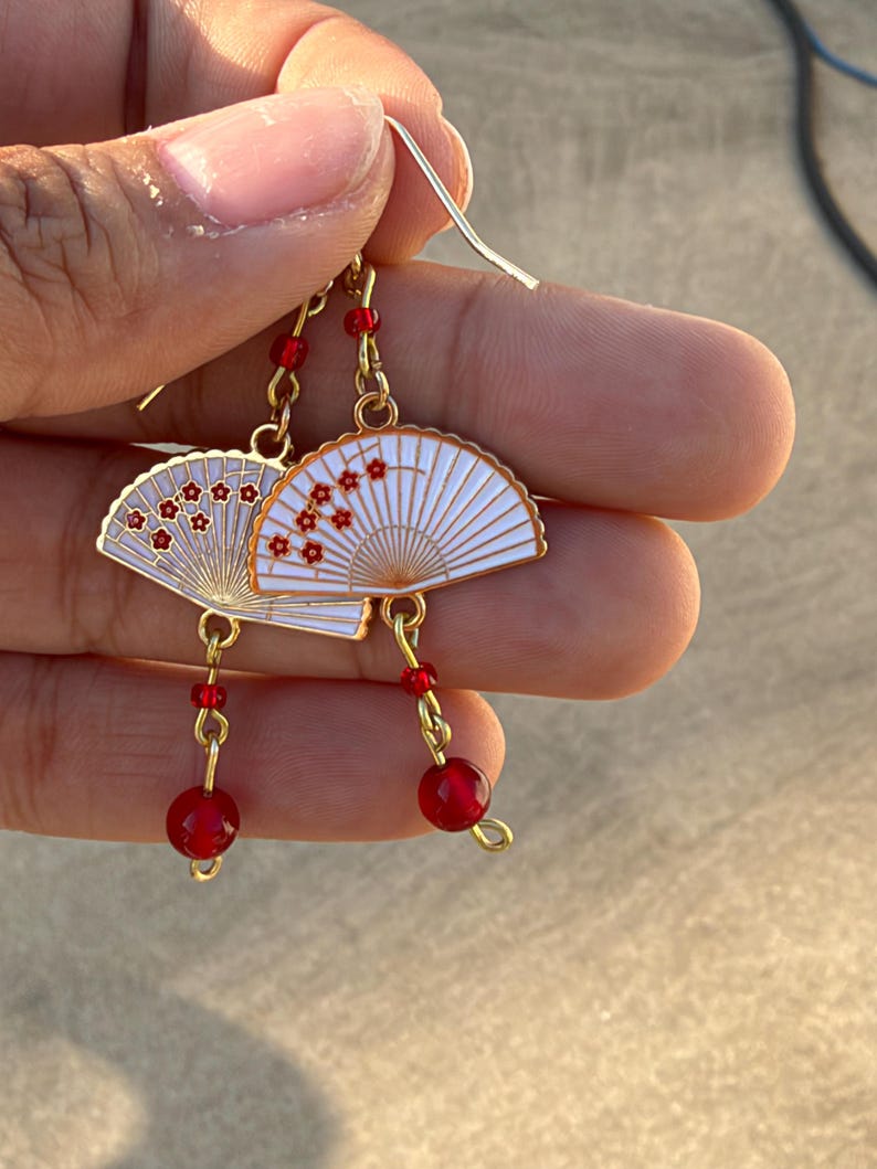May include: A pair of gold-toned dangle earrings featuring white fan-shaped charms decorated with small red floral patterns. Each earring is accented with a single small red bead above the fan and a larger red bead hanging from the bottom.