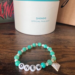 Puede incluir: Un brazalete de cuentas verdes y blancas con la palabra "ONEW" escrita en letras blancas. El brazalete tiene un pequeño dije de diamante.