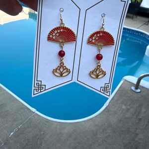 May include: A pair of gold-toned dangle earrings featuring a red fan-shaped charm with white floral details, a small red bead, and a gold lotus flower charm. The earrings are displayed on a white card with the text Thank you for supporting my business.