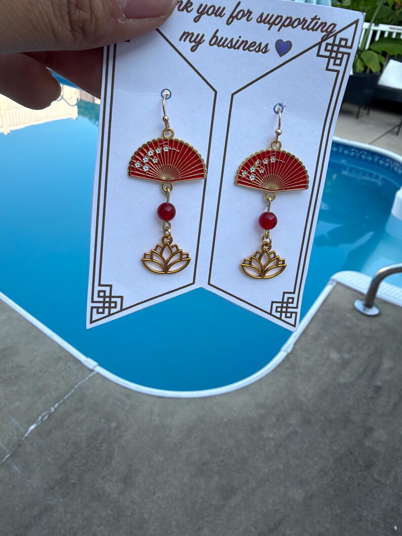 May include: A pair of gold-toned dangle earrings featuring a red fan-shaped charm with white floral details, a small red bead, and a gold lotus flower charm. The earrings are displayed on a white card with the text Thank you for supporting my business.