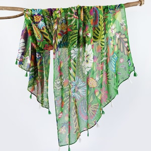 Puede incluir: Una bufanda verde transparente con estampado floral colorido y borlas blancas. La bufanda est&aacute; drapeada sobre una rama.