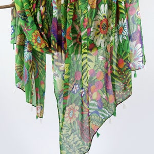 Puede incluir: Una bufanda de gasa verde con estampado floral de colores. La bufanda tiene borlas en los extremos.