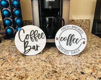 May include: Two white round signs with black lettering. The sign on the left says "Coffee Bar" with a woodgrain background. The sign on the right says "Fresh Brewed Coffee Served Daily" with coffee bean accents.
