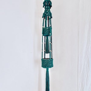 May include: A teal macrame plant hanger with three tiers and a beaded tassel. The hanger is made of cotton cord and has a wooden bead at the top.
