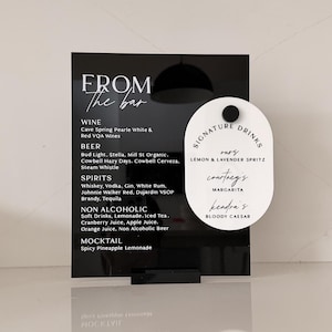 Peut inclure: Un panneau rectangulaire en acrylique noir avec du texte blanc intitulé FROM THE BAR, listant vins, bières, spiritueux, boissons sans alcool et mocktails. Un petit panneau ovale blanc intitulé SIGNATURE DRINKS est fixé dessus.