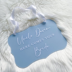 May include: A light blue acrylic sign with a white ribbon hanging from the top. The sign reads "Uncle Devin, Here Comes Your Bride" in white cursive lettering.