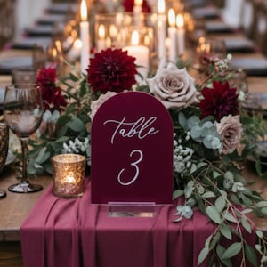 May include: A long wooden table set for a formal event, adorned with burgundy table runners, floral arrangements, and candles. A burgundy table number 3 is displayed. The table is set with black plates, gold cutlery, and wine glasses.