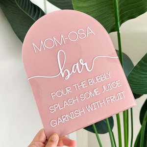 May include: A pink acrylic sign with rounded top edges. The sign reads "MOM-OSA bar" in white script. Below, the sign lists instructions: "Pour the bubbly, splash some juice, garnish with fruit."
