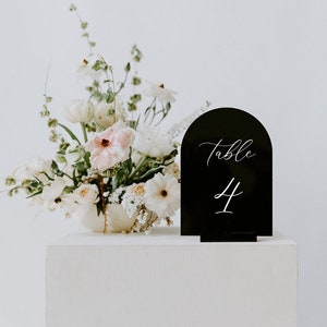May include: Black acrylic table number with a rounded arch top. The number "4" is written in white script on the sign. The sign is sitting on a white pedestal in front of a white wall. A white and pink floral arrangement is in the background.