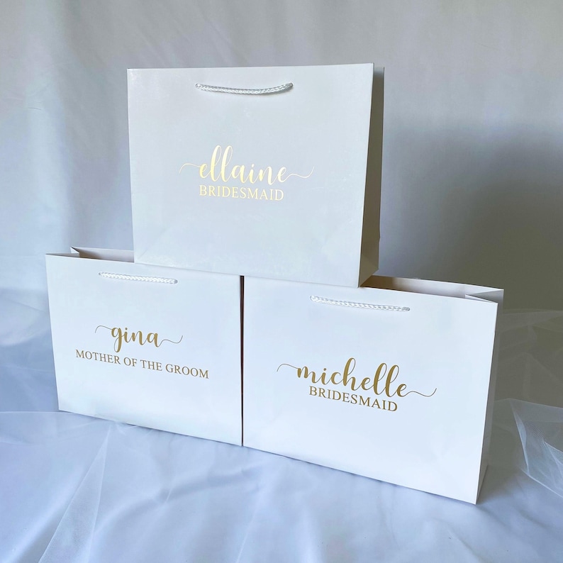 May include: Three white gift bags with gold lettering. The top bag says "ellaine bridesmaid". The middle bag says "gina mother of the groom". The bottom bag says "michelle bridesmaid".