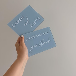 May include: Two rectangular dusty blue acrylic signs with white lettering. The top sign features the text CARDS and GIFTS. The bottom sign features the text PLEASE SIGN OUR guestbook. The signs have a smooth, matte finish and clean edges.