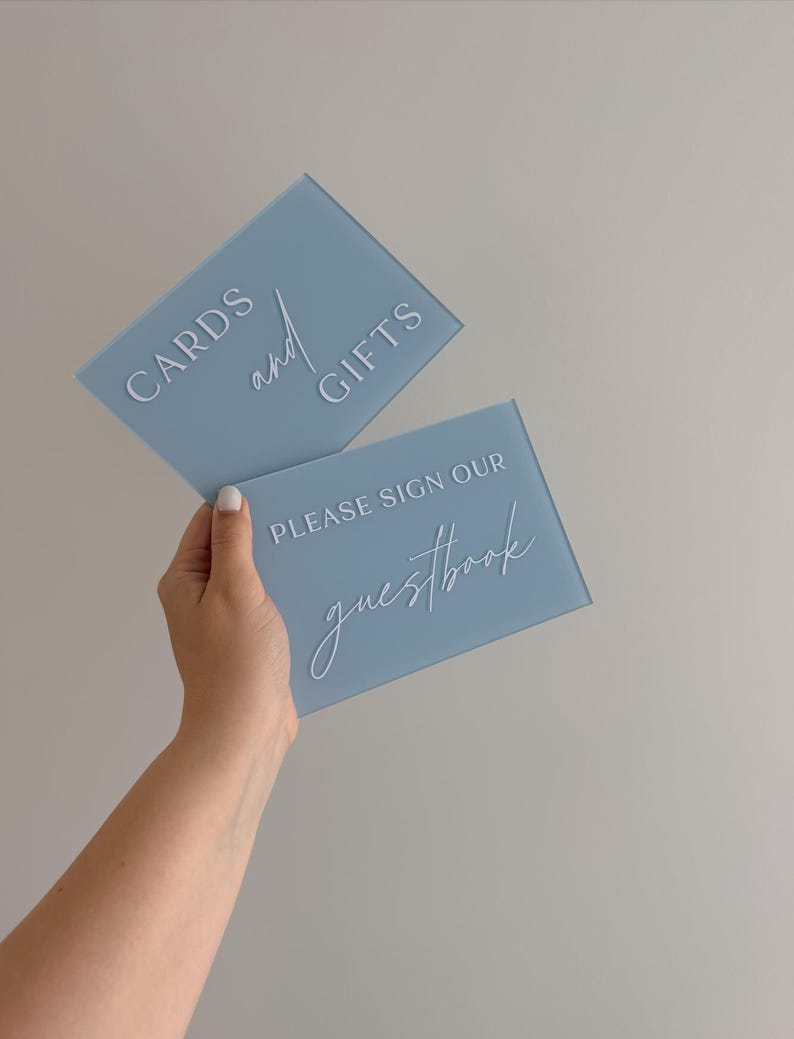 May include: Two rectangular dusty blue acrylic signs with white lettering. The top sign features the text CARDS and GIFTS. The bottom sign features the text PLEASE SIGN OUR guestbook. The signs have a smooth, matte finish and clean edges.