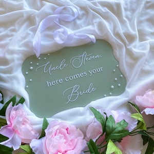 May include: Sage green wedding sign with the text "Uncle Steven, here comes your Bride" in elegant white script. The sign is adorned with small pearl-like embellishments and a white ribbon. Pink peonies and greenery surround the sign.