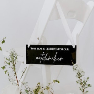 May include: Black sign with white text that reads "This seat is reserved for our matchmaker". The sign is attached to a white chair.