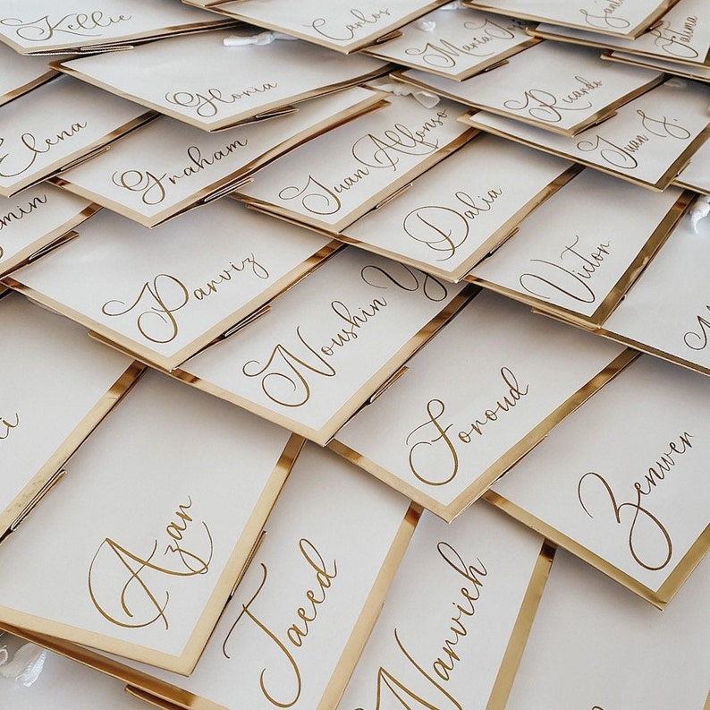 May include: A close-up of a pile of white place cards with gold lettering. The cards are arranged in a random pattern and have a gold border. The names on the cards include "Azan", "Nowshen", "Foroud", "Benven", "Jalia", "Vitor", "Parviz", "Graham", "Gloria", "Juan Alonso", "Taheed", "Manvich", "Kelly", "Lena", and "Carlos".