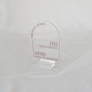 May include: Clear acrylic sign with the text "signature drinks" in a curved arc at the top. Below the text, the sign reads "HIS OLD FASHION" and "HERS MARILYN MONROE".