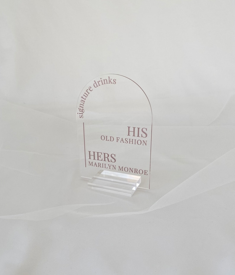 May include: Clear acrylic sign with the text "signature drinks" in a curved arc at the top. Below the text, the sign reads "HIS OLD FASHION" and "HERS MARILYN MONROE".