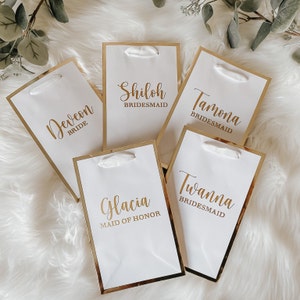May include: Five white gift bags with gold trim and white ribbon handles. The bags are labeled with the names "Deveon Bride", "Shiloh Bridesmaid", "Tamona Bridesmaid", "Glacia Maid of Honor", and "Twanna Bridesmaid".