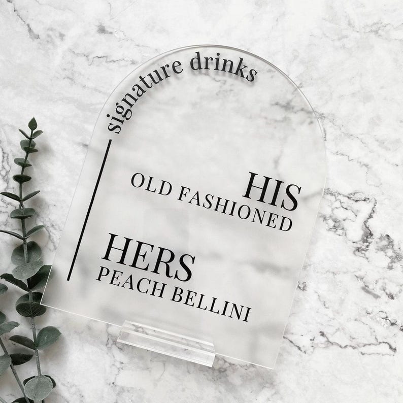 May include: Clear acrylic sign with the text "signature drinks" in a curved arch at the top. Below the text, it says "His Old Fashioned" and "Hers Peach Bellini".
