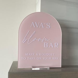 May include: A pink acrylic sign with rounded edges that reads "AVA'S bloom BAR MAKE A BOUQUET TO TAKE ON YOUR WAY".