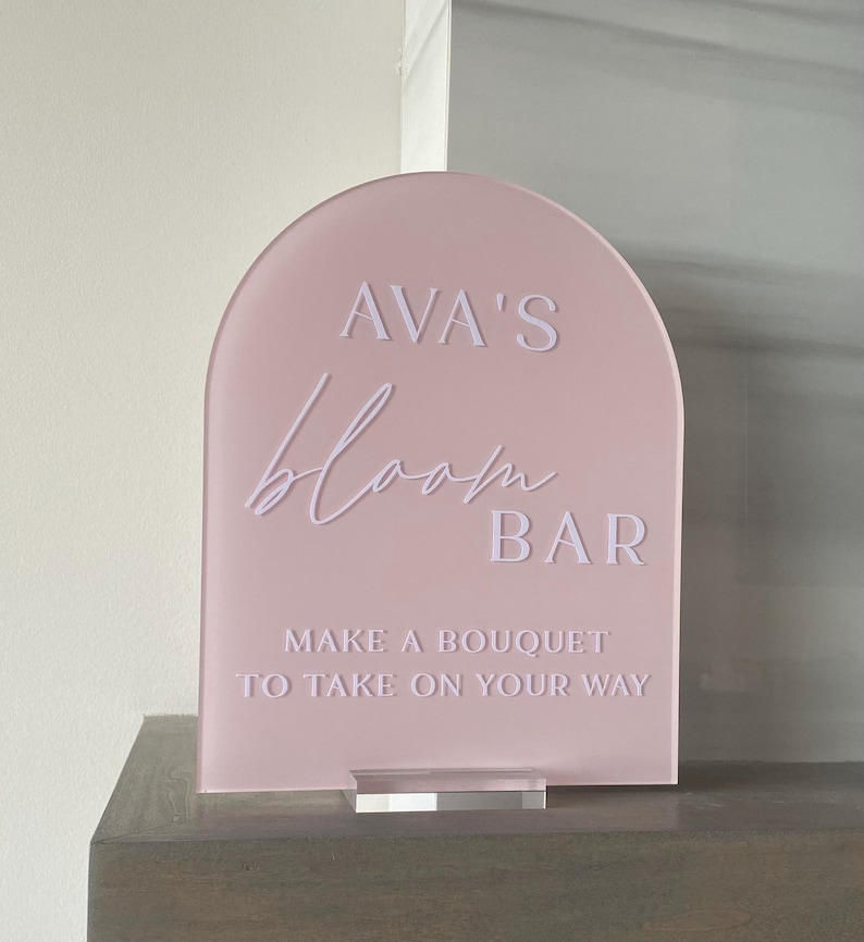 May include: A pink acrylic sign with rounded edges that reads "AVA'S bloom BAR MAKE A BOUQUET TO TAKE ON YOUR WAY".