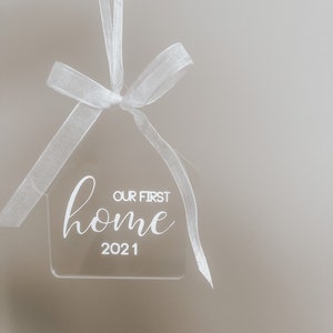 Puede incluir: Adorno de casa de acr&iacute;lico blanco con el texto "Our First Home 2021" colgado de una cinta blanca.