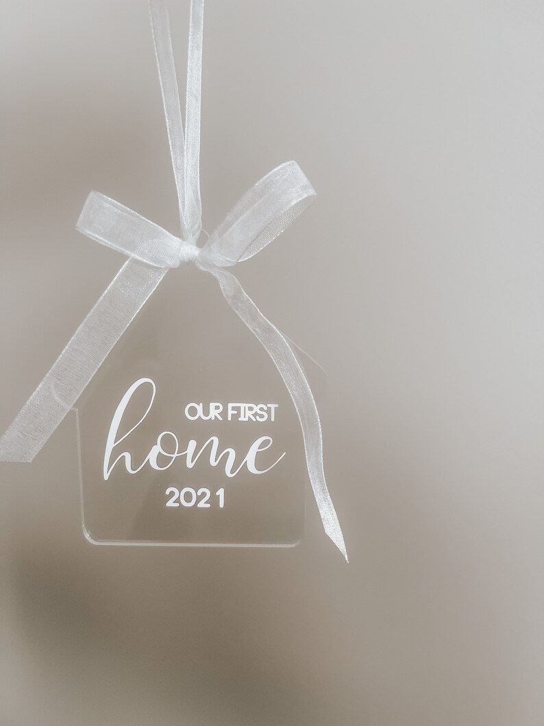 Puede incluir: Adorno de casa de acr&iacute;lico blanco con el texto "Our First Home 2021" colgado de una cinta blanca.