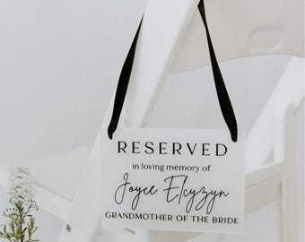 May include: White chair with a black ribbon and a white sign that says "RESERVED in loving memory of Joyce Elyzyn GRANDMOTHER OF THE BRIDE"