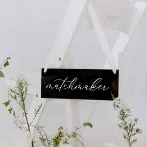 May include: Black acrylic sign with white calligraphy that reads "matchmaker" hanging on a white chair.