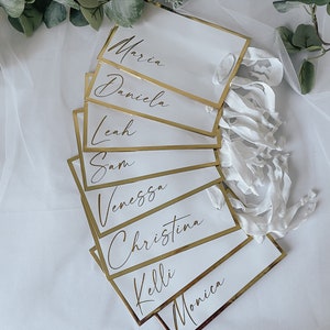 May include: White place cards with gold trim and handwritten names, including Maria, Daniela, Leah, Sam, Vanessa, Christina, Kelli, and Monica.