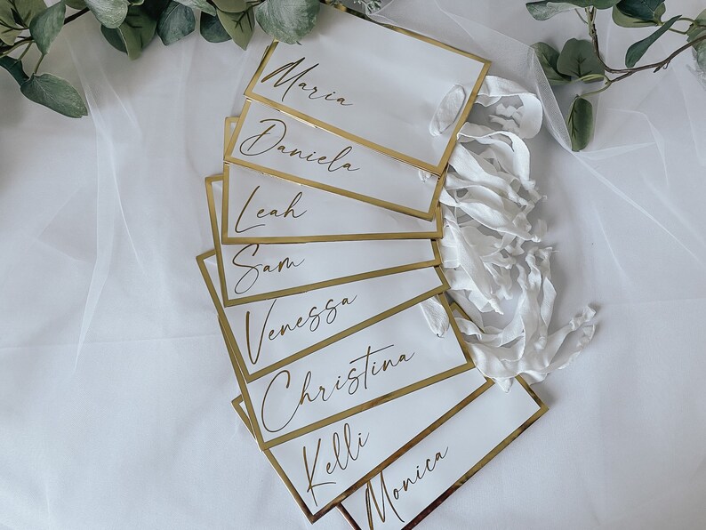 May include: White place cards with gold trim and handwritten names, including Maria, Daniela, Leah, Sam, Vanessa, Christina, Kelli, and Monica.