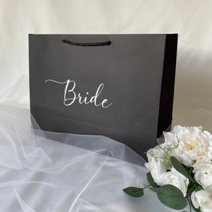 May include: A black gift bag with a white "Bride" inscription. The bag is sitting on a white tulle fabric with a white flower bouquet.