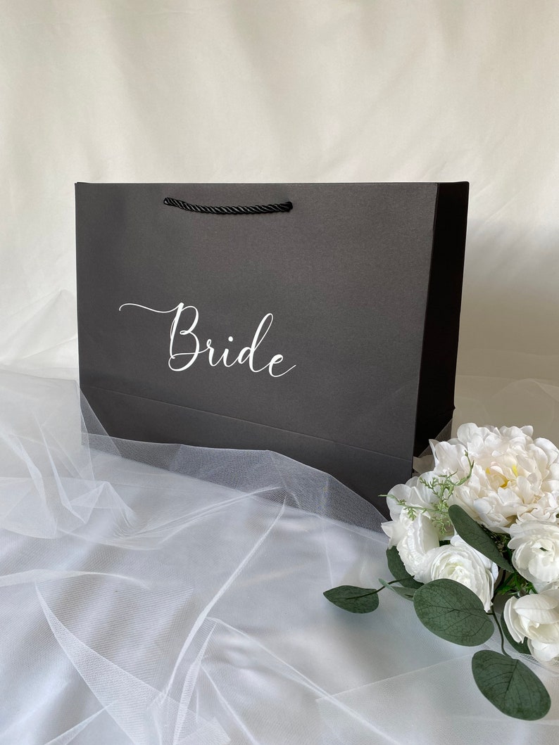 May include: A black gift bag with a white "Bride" inscription. The bag is sitting on a white tulle fabric with a white flower bouquet.