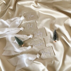 May include: Five clear acrylic gift tags with white lettering. The tags are tied with white ribbon and have the names "Mom", "Dad", "Oakley", "Bourbon", and "Brody" written on them.